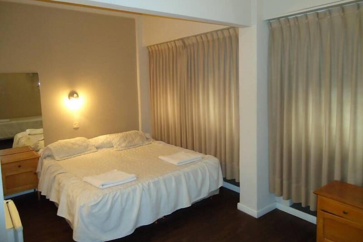 Un lit bien fait, avec du linge de lit blanc, une table de chevet et une lampe fixée au mur.