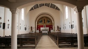 An interior of a church with rows of wooden pews, a central altar, and a red and gold stained glass window.
