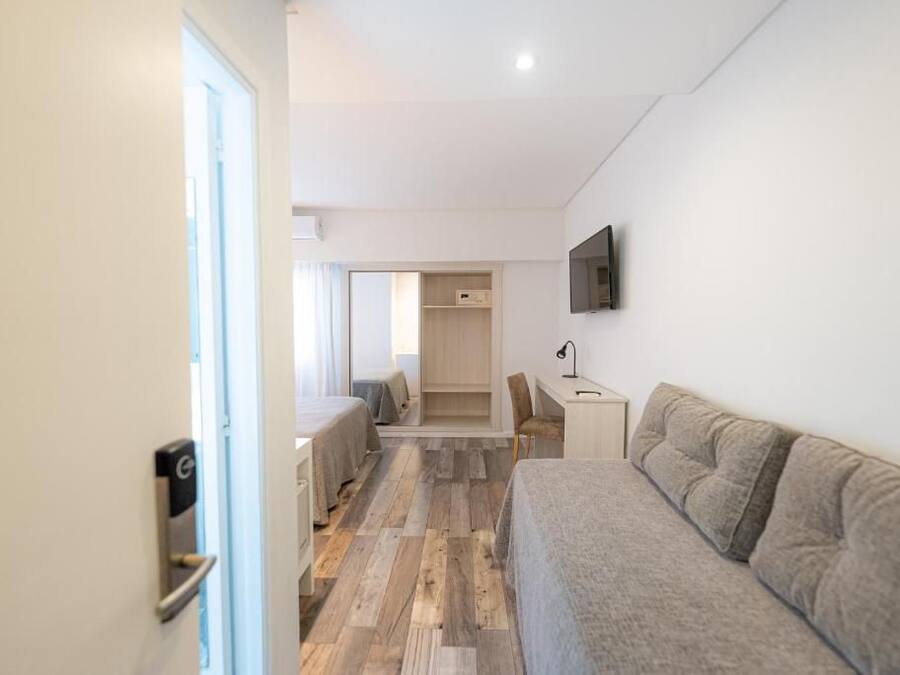 A modern hotel room with a wooden floor, a sofa, a desk, a chair, a TV mounted on the wall, and a door with a handle.