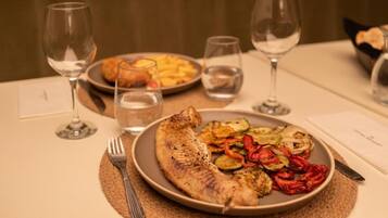 Una mesa de comedor con un plato de pescado a la parrilla y verduras, copas de vino y una cesta de pan.