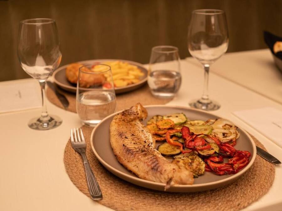 Una mesa de comedor con un plato de pescado a la parrilla y verduras, copas de vino y una cesta de pan.