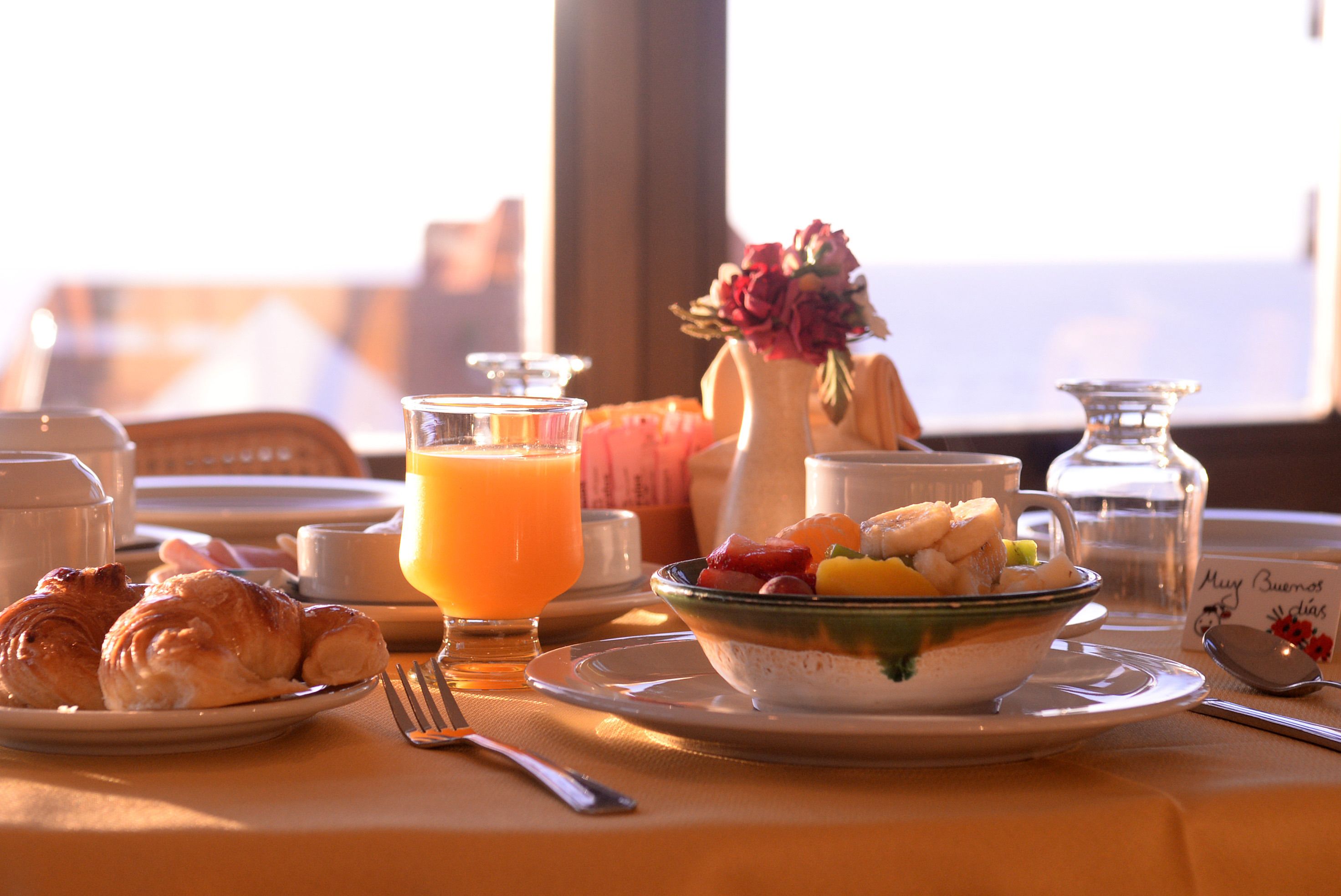 Una mesa de desayuno con croissants, un vaso de jugo de naranja, un bol de fruta y un jarrón con flores.