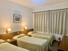 Habitación de hotel con dos camas, cabecero de madera, mesita de noche con lámpara, un cuadro enmarcado en la pared y una ventana con cortinas.