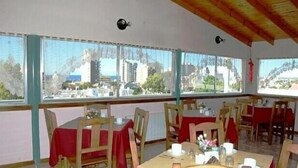 A dining area with tables set for four, wooden chairs, and a view of a cityscape through large windows. - Hotel Fantilli (Puerto Madryn)