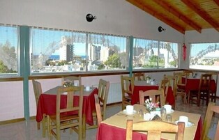 Comedor con mesas dispuestas para cuatro, sillas de madera y vistas a un paisaje urbano a través de amplias ventanas.