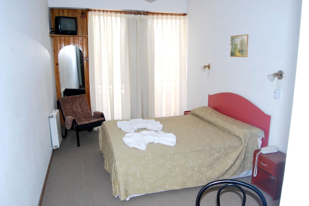 A hotel room with a bed, a chair, a radiator, a television, and a bedside table with a lamp.