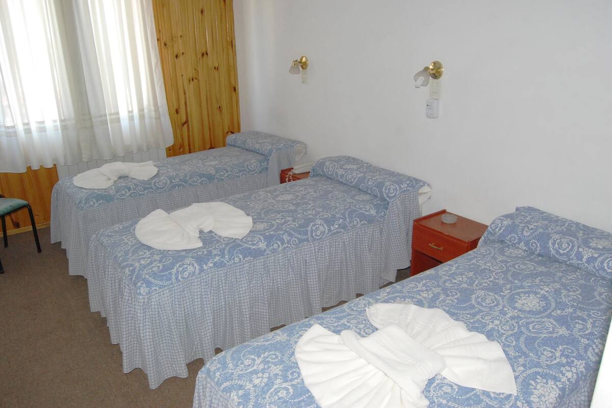 A room with three single beds, each with a blue patterned bedspread and white towels. There is a wooden door and a small wooden nightstand with a red drawer.