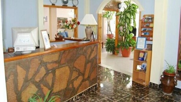A reception area with a stone counter, a desk with a computer, a lamp, and a bookshelf with books and a potted plant. - Hotel Fantilli (Puerto Madryn)