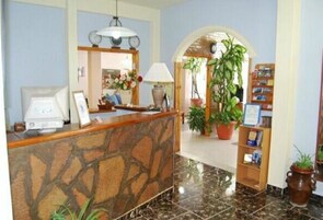 Una zona de recepción con mostrador de piedra, un escritorio con computadora, una lámpara y una estantería con libros y una planta en maceta.