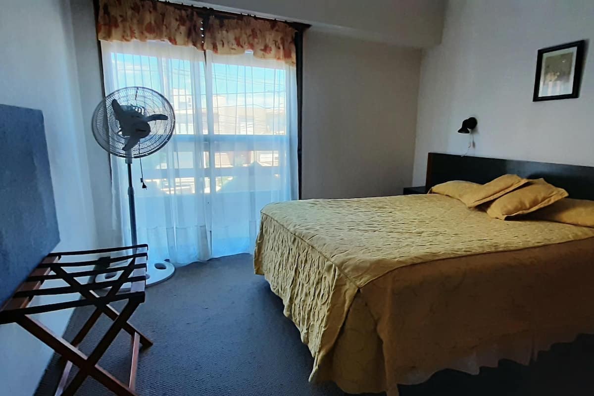 Une chambre à coucher avec un lit, un ventilateur sur pied, une fenêtre avec des rideaux et un tableau accroché au mur.