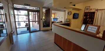 Zona de recepción de un hotel, con mostrador, un reloj y un cartel enmarcado que indica el horario de administración.