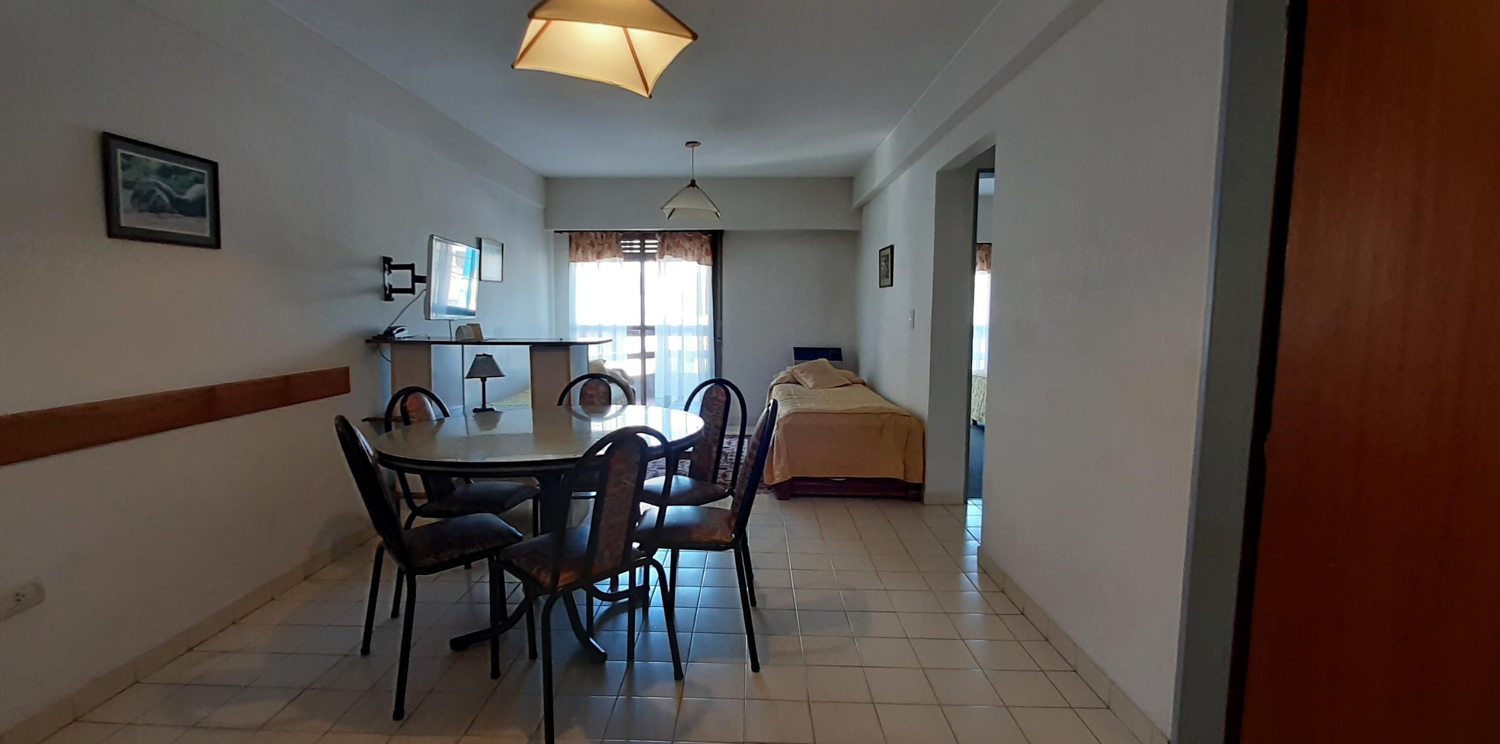 Habitación funcional con una mesa de comedor y sillas, una cama, un escritorio con computadora y una ventana con cortinas.