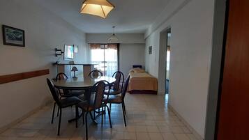 Habitación funcional con una mesa de comedor y sillas, una cama, un escritorio con computadora y una ventana con cortinas.