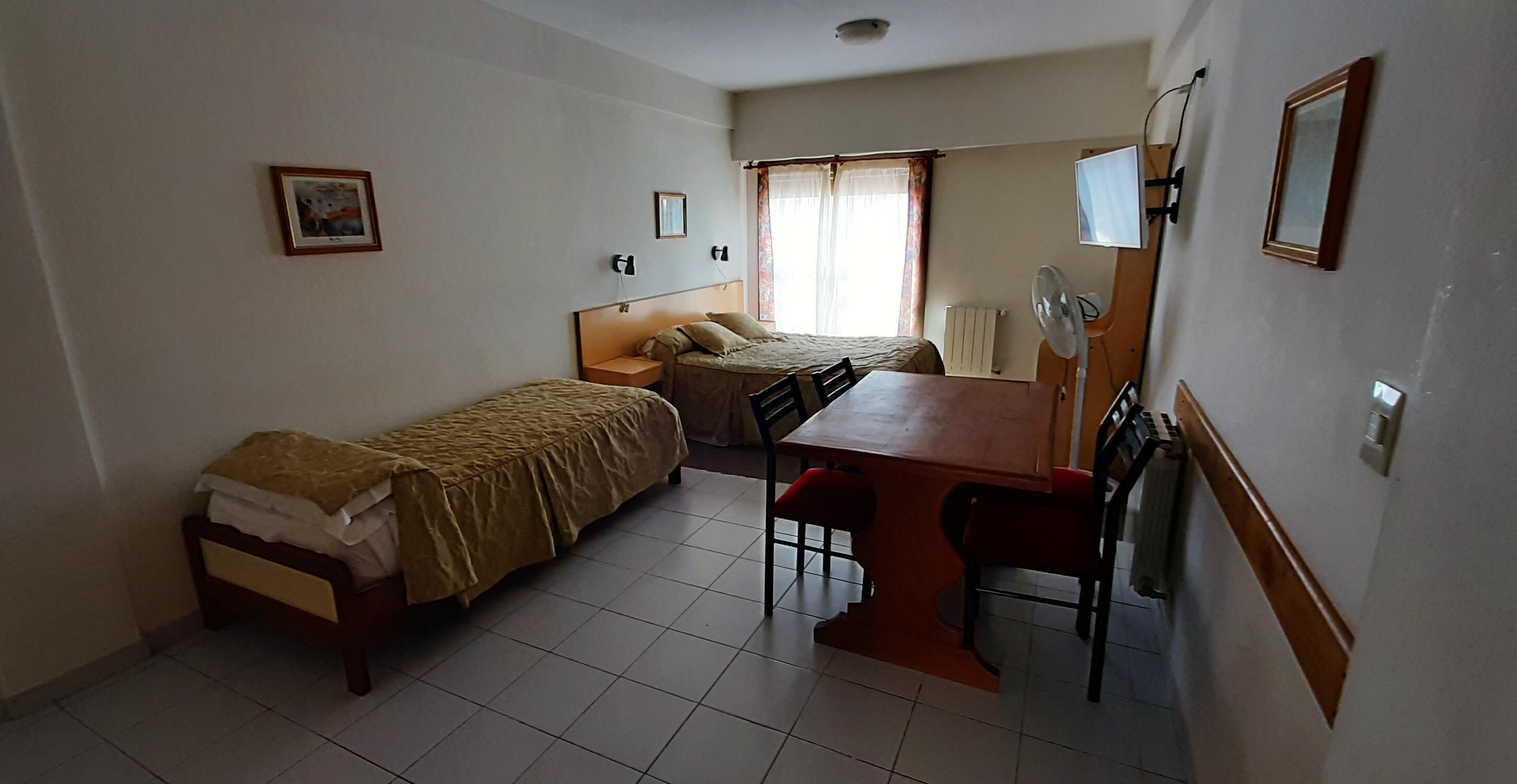 A compact hotel room with two single beds, a small table with chairs, a desk, a chair, a mirror, and a radiator.