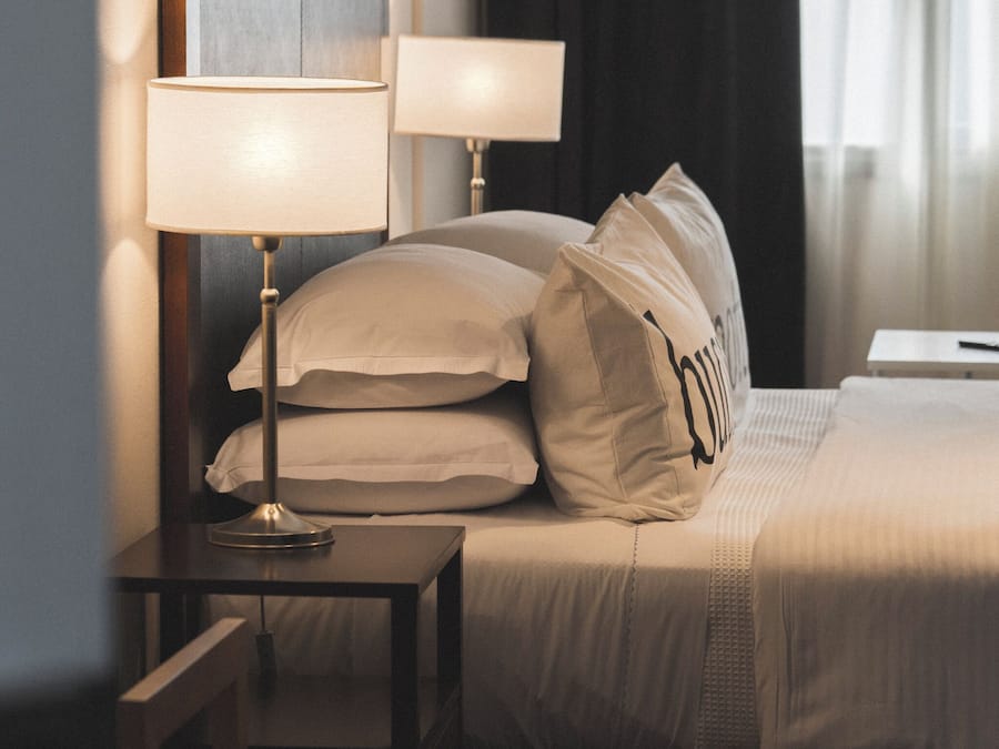 A neatly made bed with pillows, a bedside table with a lamp, and a dark curtain in a hotel room.
