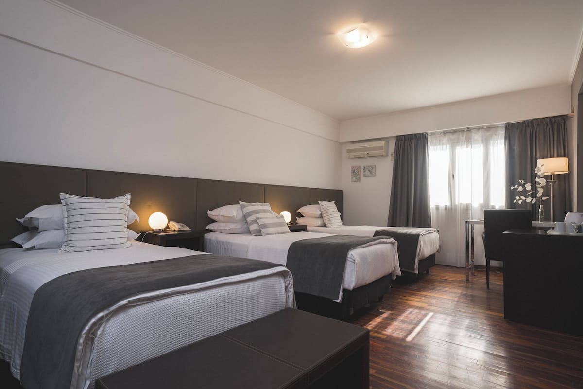 Une chambre d’hôtel avec deux lits, un bureau, une chaise, une lampe et une fenêtre avec des rideaux.