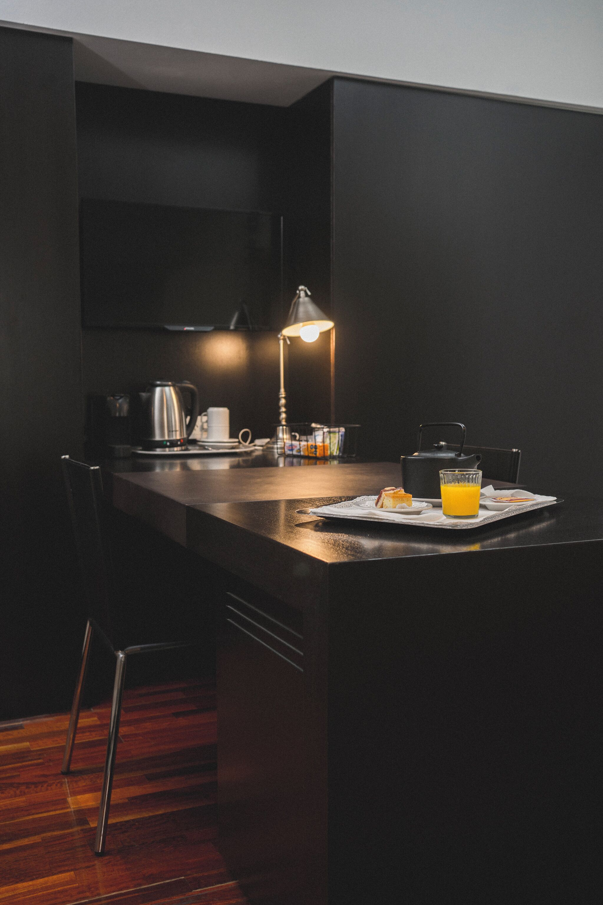 Une cuisine moderne avec un plan de travail foncé, une bouilloire, une machine à café, un verre de jus d’orange et une lampe.