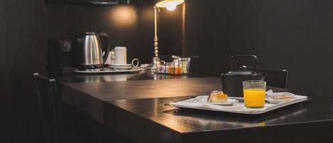 Une cuisine moderne avec un plan de travail foncé, une bouilloire, une machine à café, un verre de jus d’orange et une lampe.