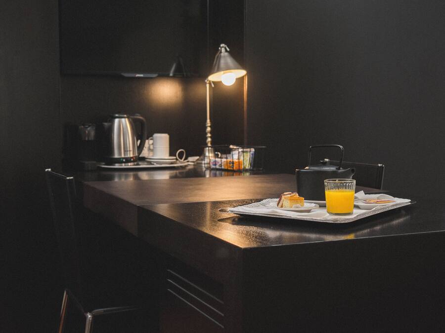 Una cocina moderna con encimera oscura, una tetera, una cafetera, un vaso de jugo de naranja y una lámpara.