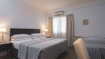 Habitación de hotel con cama doble, cama individual, escritorio con lámpara, una silla, aire acondicionado y radiador.