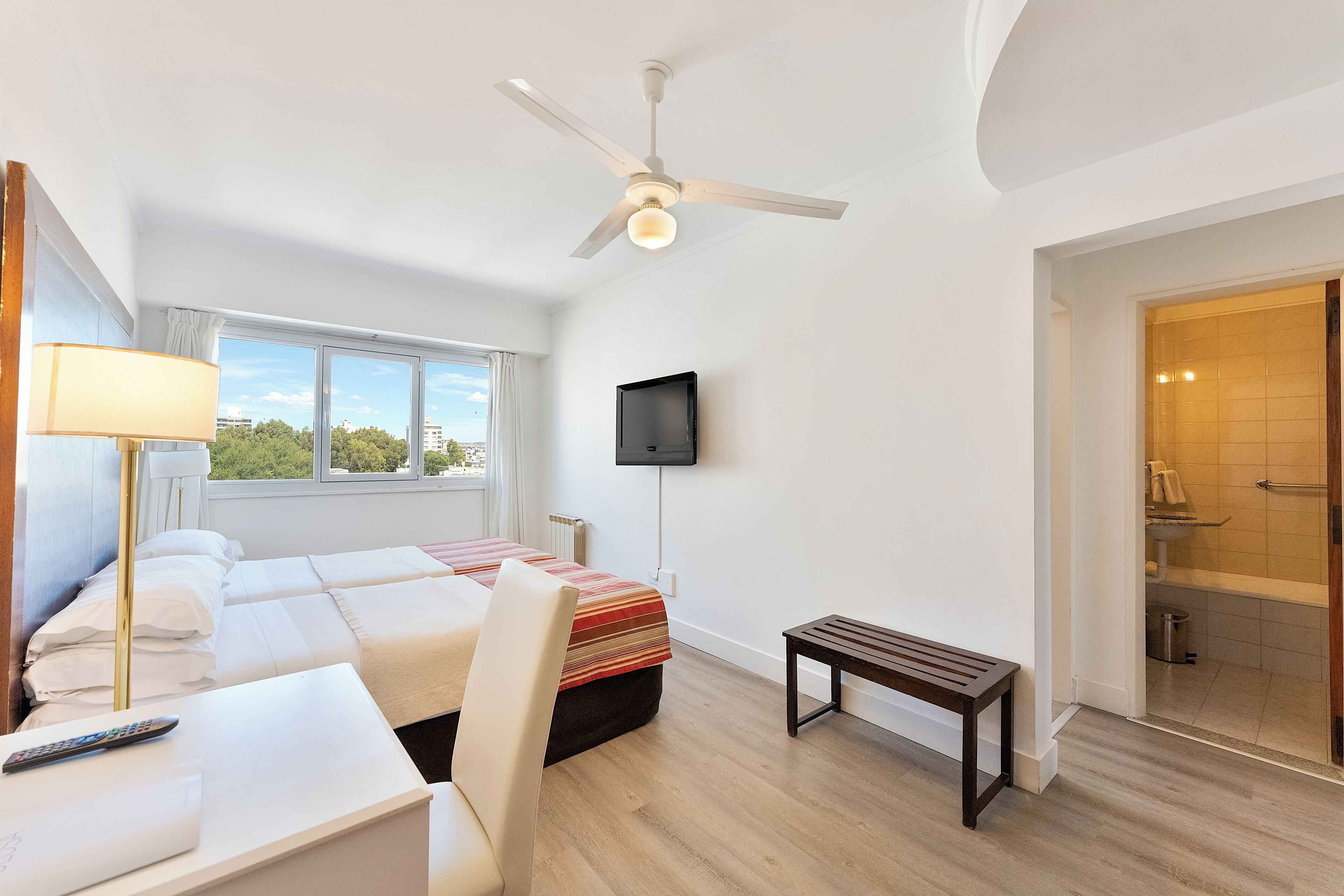 A hotel room with a bed, a desk, a chair, a bench, a ceiling fan, a TV, and a bathroom visible through an open door.