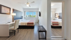 Habitación de hotel con dos camas, un escritorio, una silla, un banco, un ventilador de techo y un televisor.