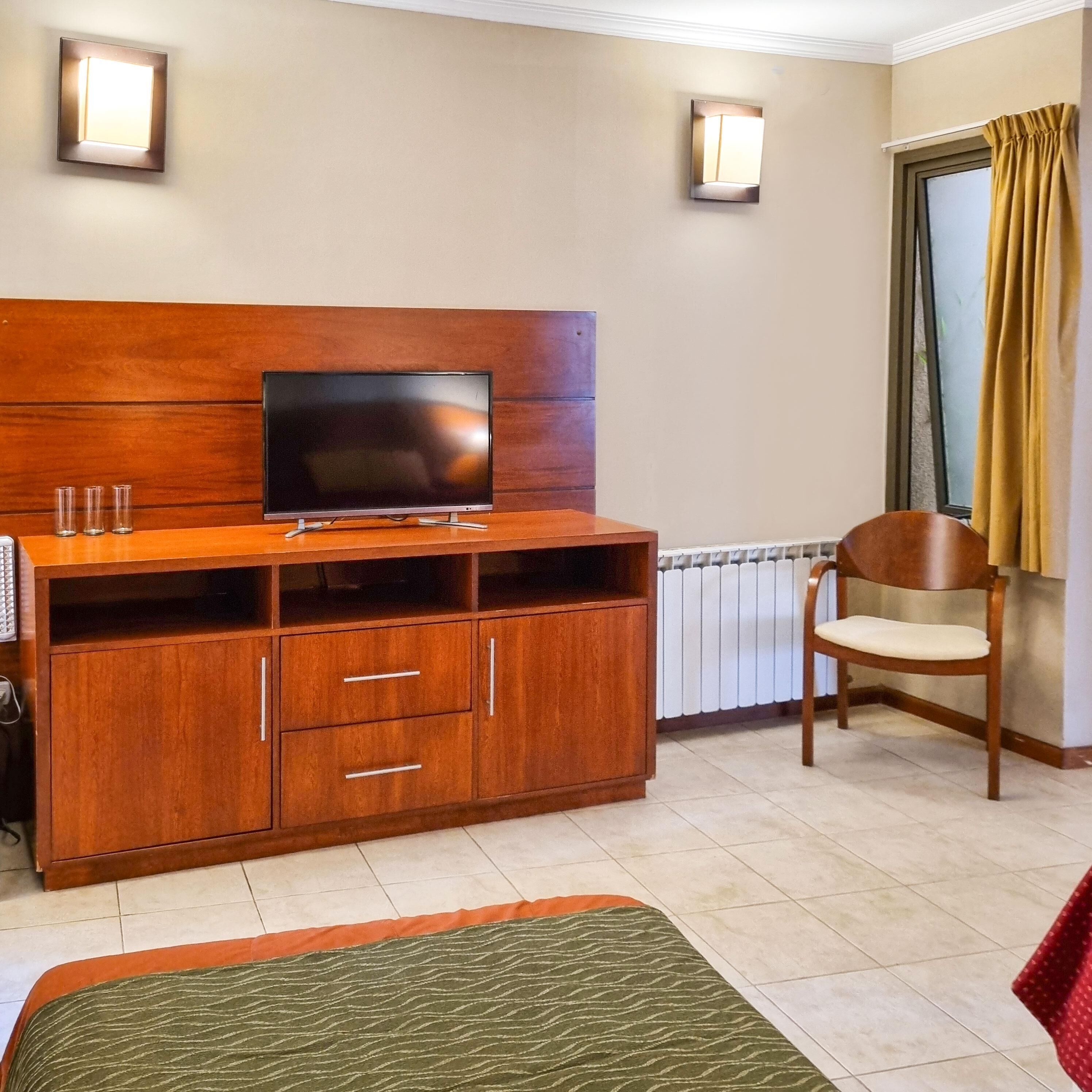 Habitación de hotel con un mueble de televisión de madera, un televisor de pantalla plana, una silla de madera, un radiador y una ventana con cortinas.