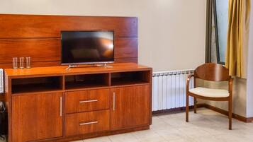 Habitación de hotel con un mueble de televisión de madera, un televisor de pantalla plana, una silla de madera, un radiador y una ventana con cortinas.