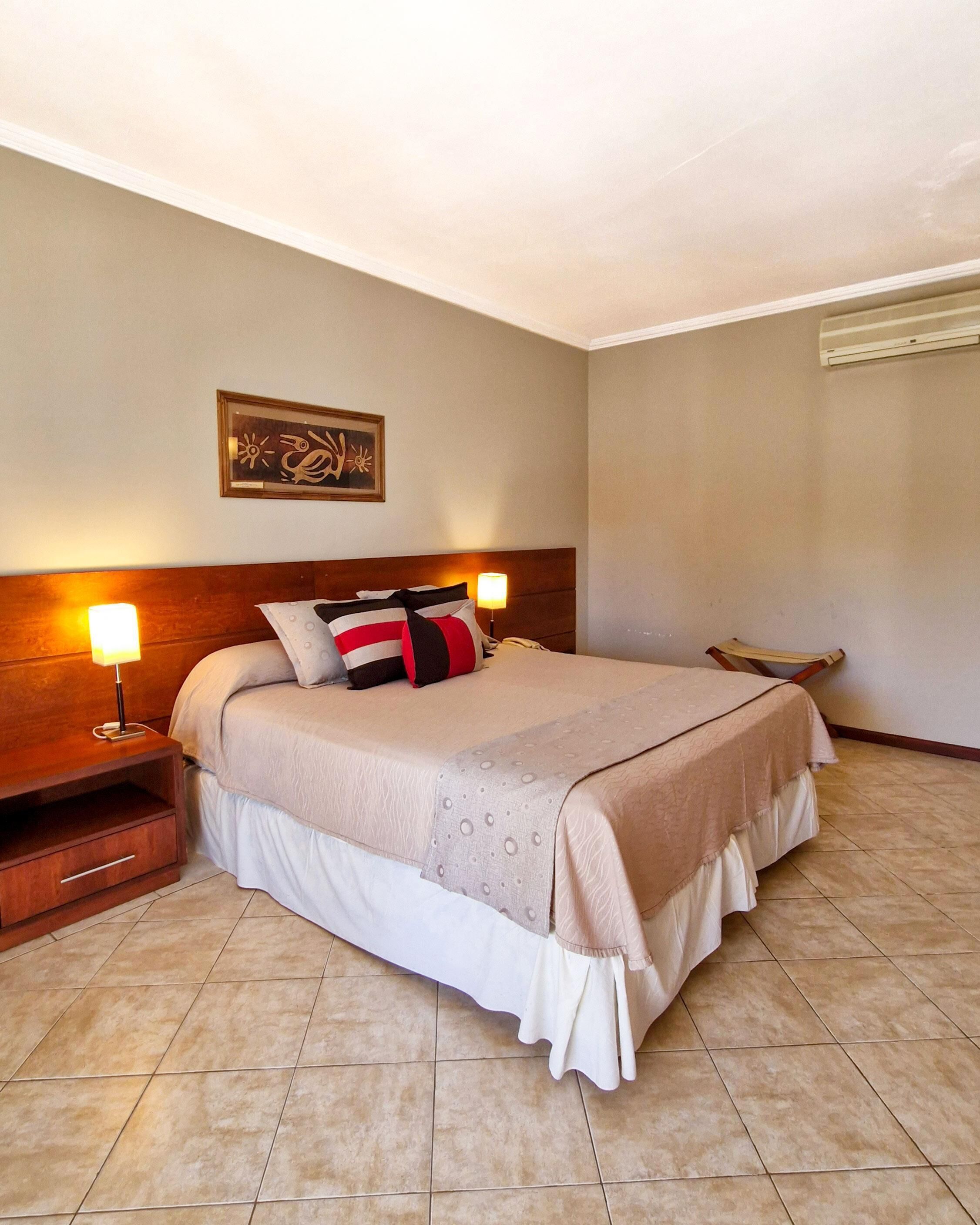 Una cama bien hecha con cabecera, una mesita de noche con lámpara y un aire acondicionado instalado en la pared.