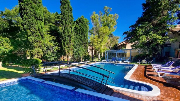 Una piscina con borde de azulejos, rodeada de árboles y una zona cubierta con sillas de descanso.
