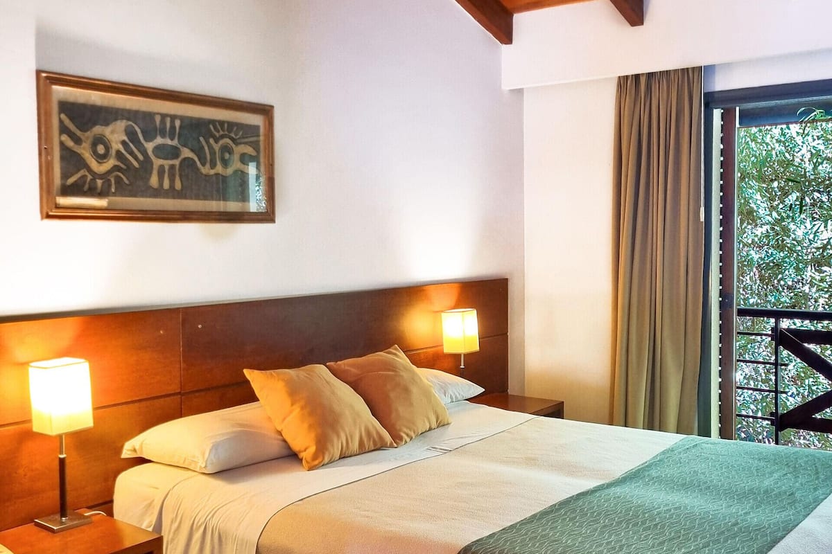 Habitación de hotel con cama, mesitas de noche, lámpara, un cuadro y vista a árboles por la ventana.