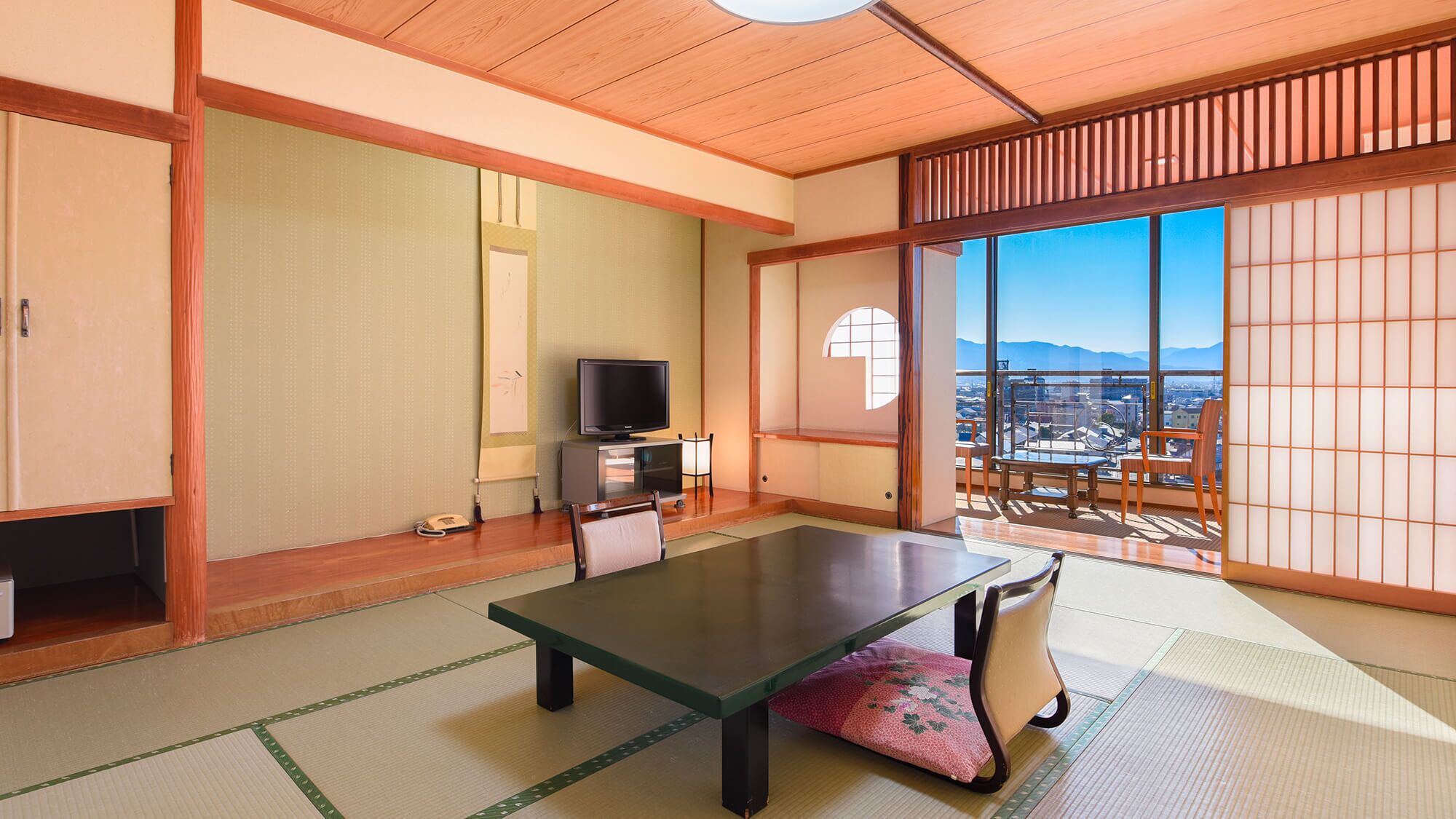 A traditional Japanese room with tatami flooring, a low table, a television, and a sliding door leading to a balcony with a city view.