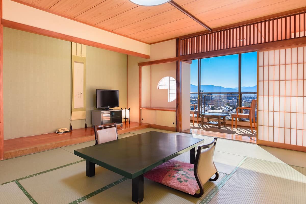 A traditional Japanese room with tatami flooring, a low table, a television, and a sliding door leading to a balcony with a city view.
