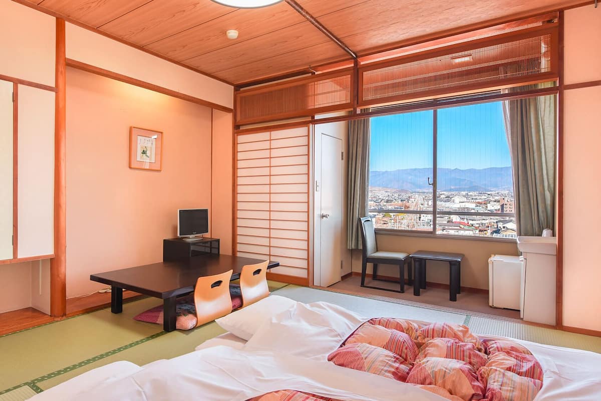 A traditional Japanese room with tatami flooring, a futon bed, a desk with a computer, and a sliding door leading to a balcony with a city view.