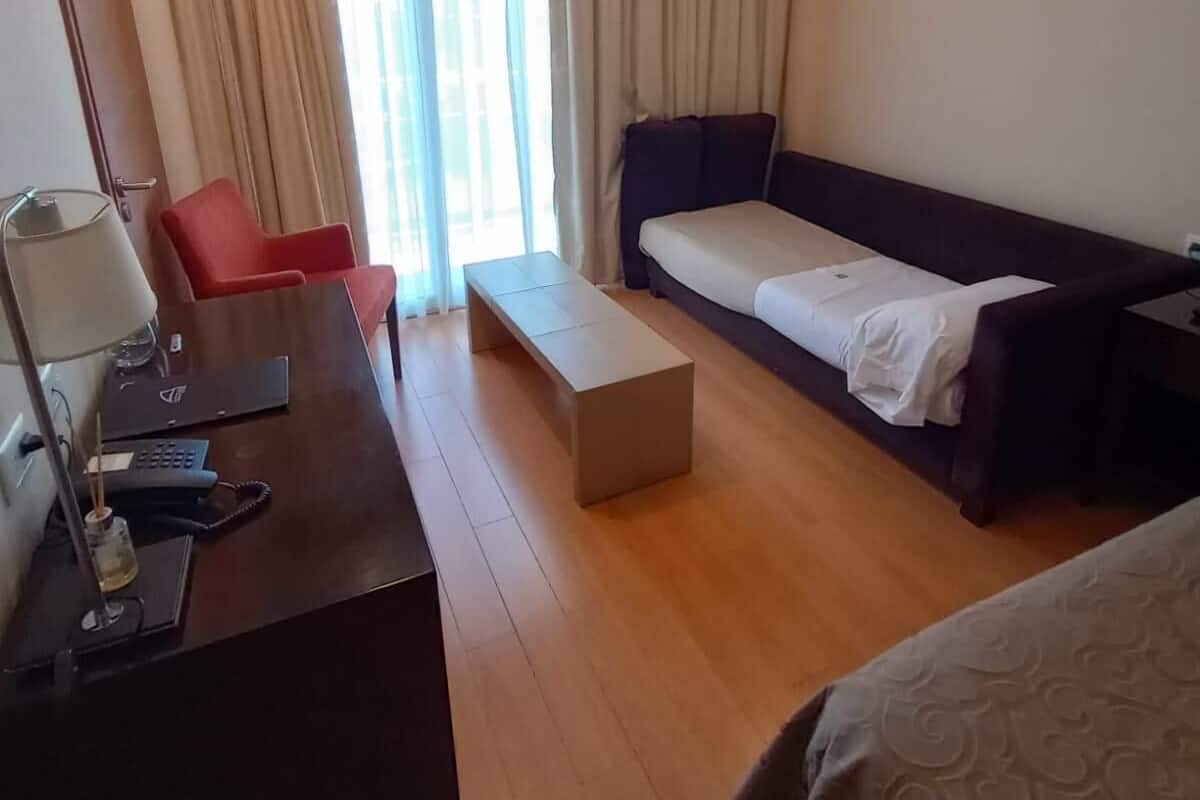 Una habitación de hotel con piso de madera, un escritorio con lámpara, una silla roja, un sofá y una cama con cubrecama estampado.