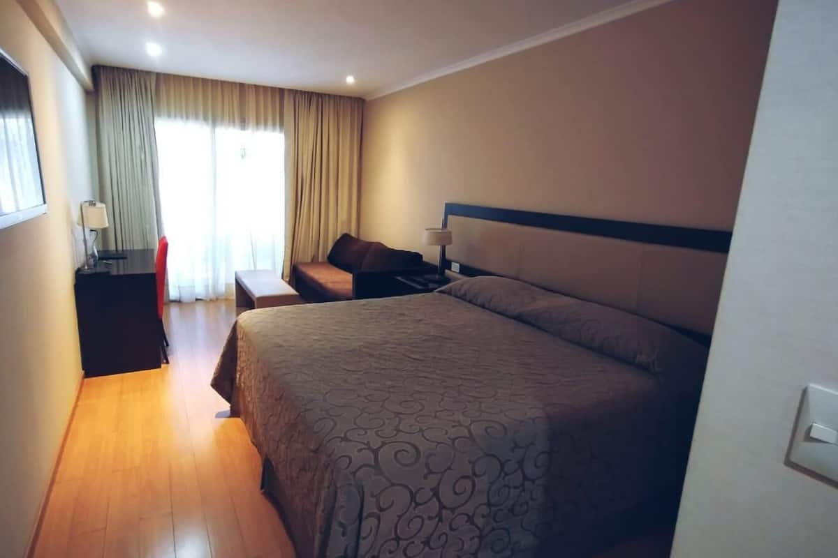 Una habitación de hotel con una cama grande, un escritorio con lámpara, una silla y un televisor montado en la pared.