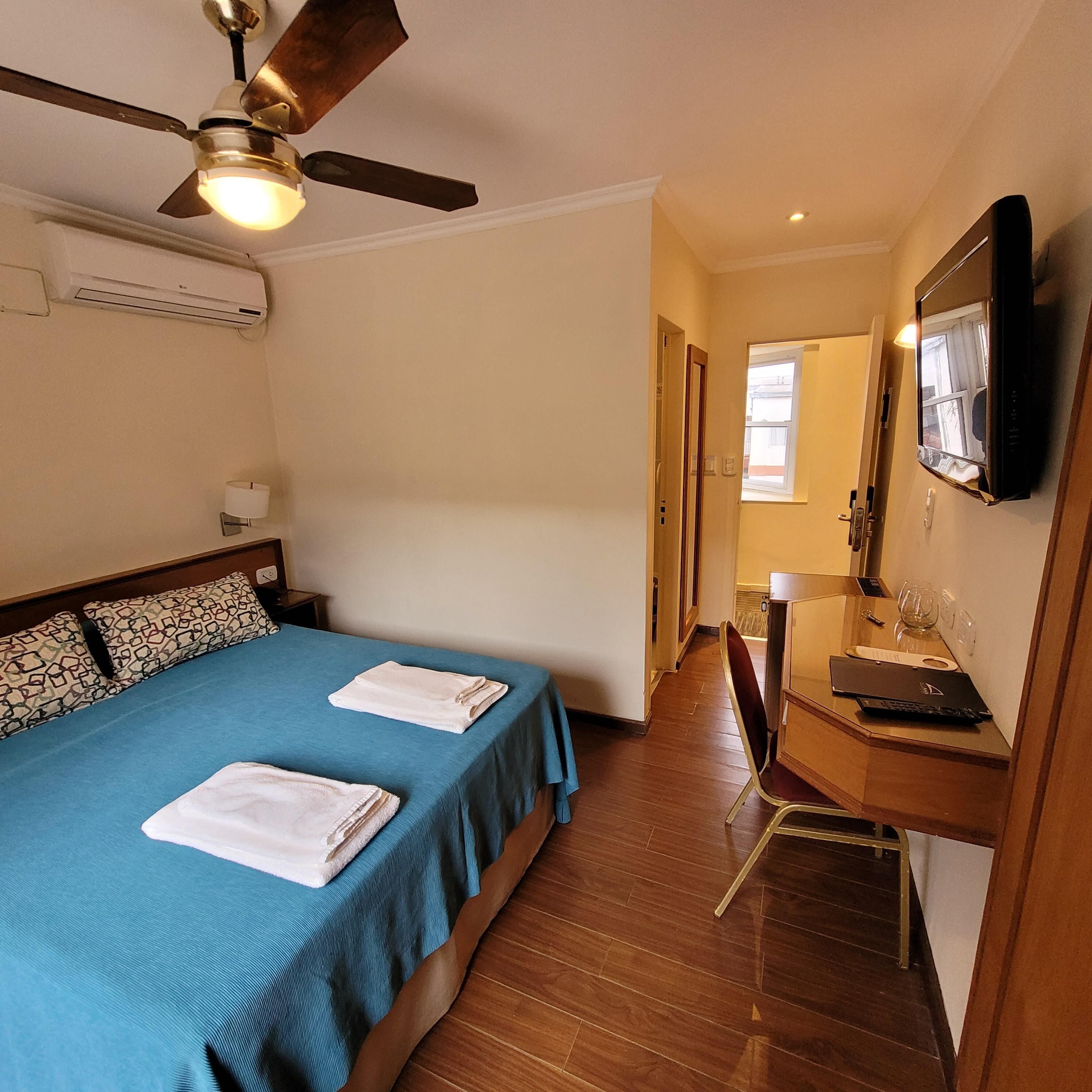 A hotel room with a bed, a desk, a chair, a television, and a ceiling fan.