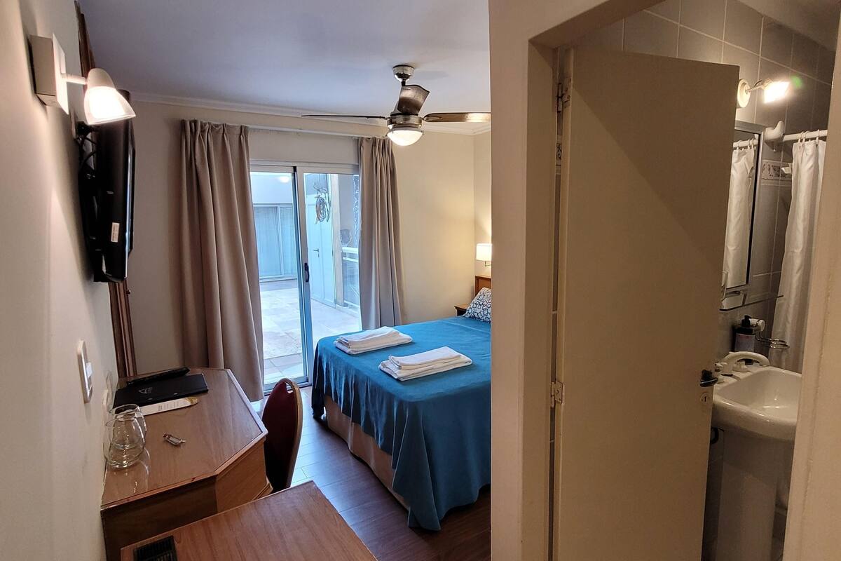 Habitación de hotel con cama, escritorio, silla, ventilador de techo, baño con lavabo y espejo, y vista al exterior.