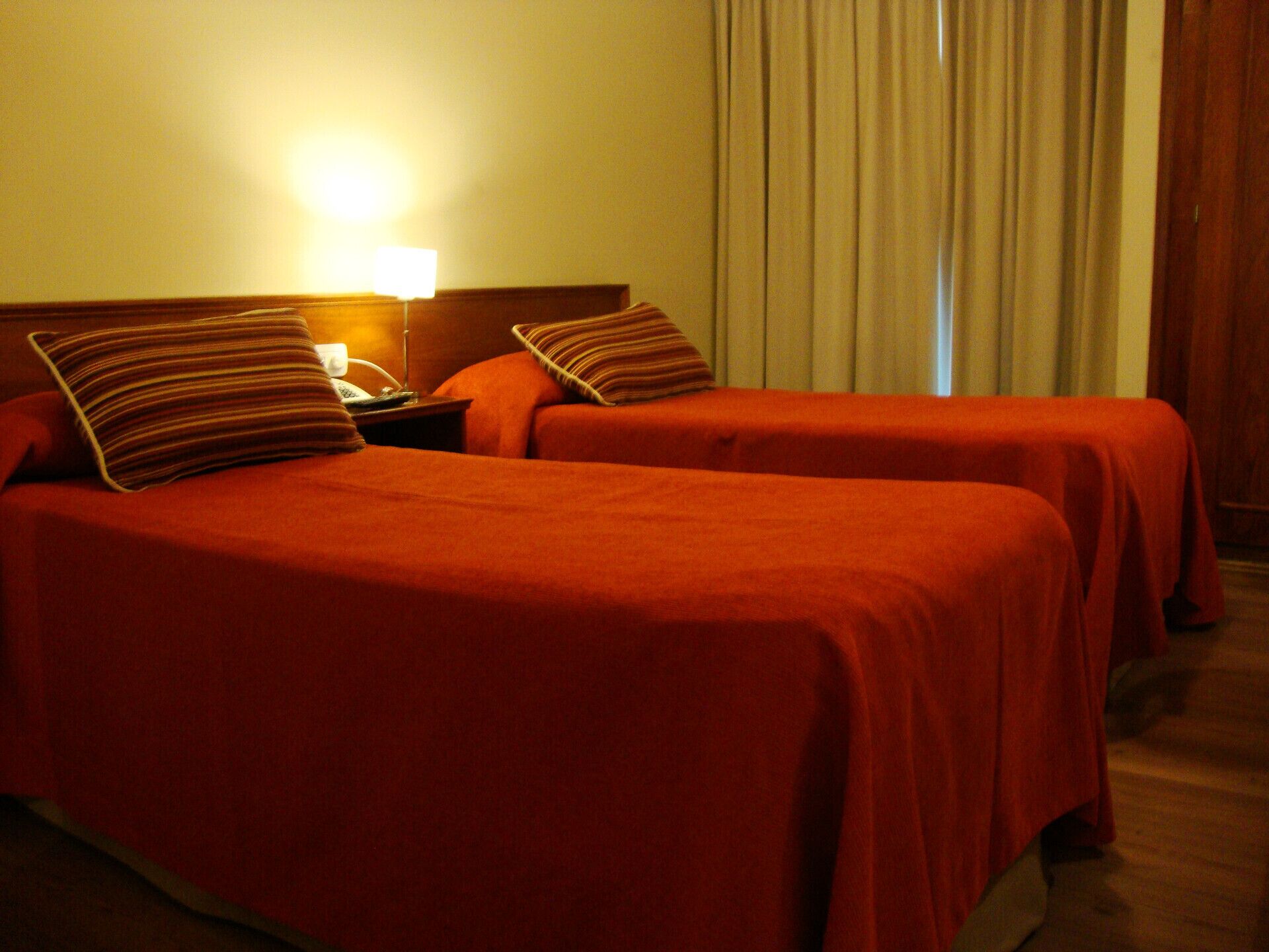 A hotel room with two beds, each with a red bedspread and striped pillows. There is a nightstand with a lamp and a telephone.