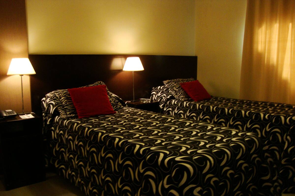 Habitación de hotel con dos camas, cada una con una colcha estampada y almohadas rojas. Hay dos mesitas de noche con lámparas, y un teléfono sobre la mesa de la izquierda.