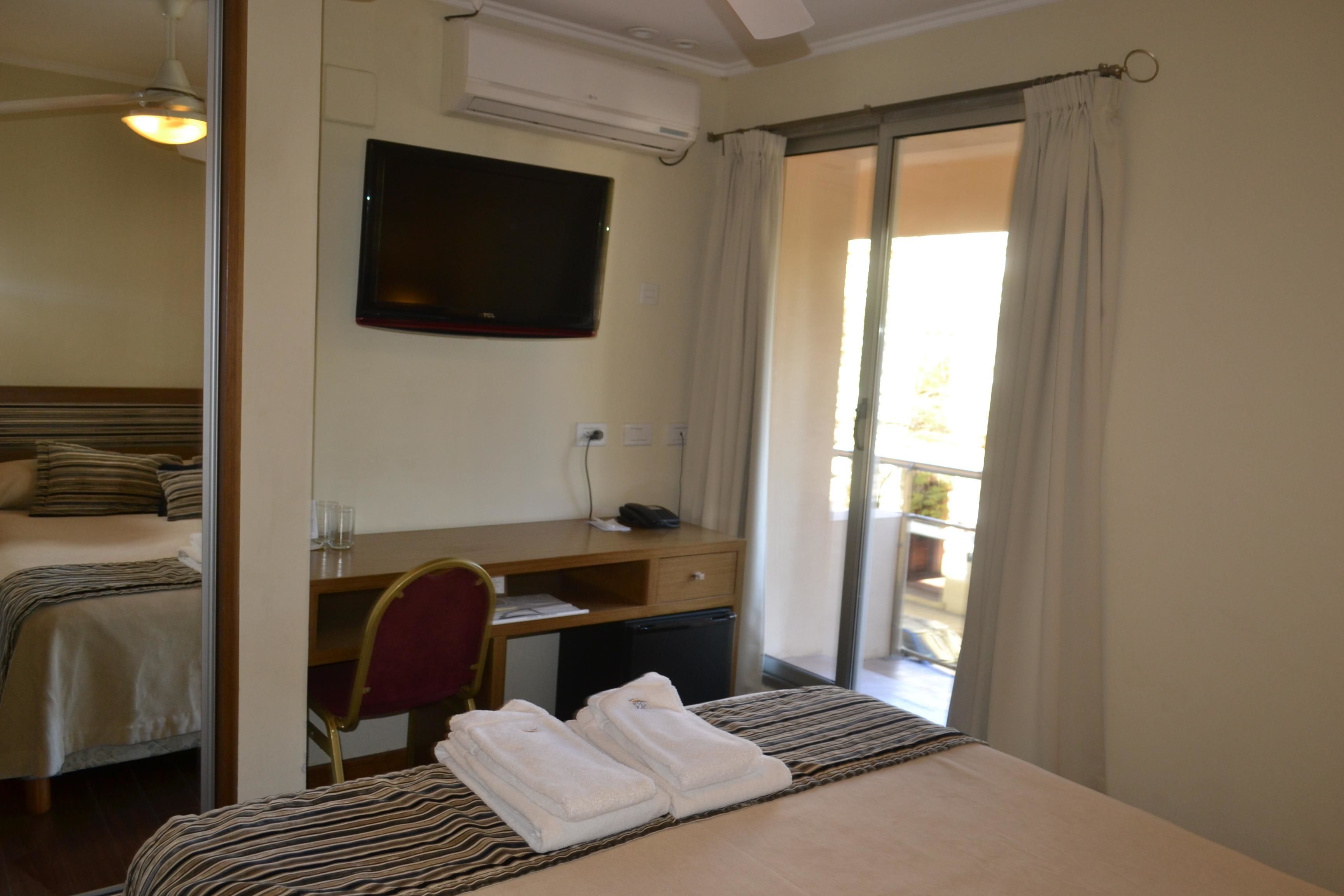 Quarto de hotel com cama, escrivaninha e cadeira, televisão e porta de correr para uma varanda.