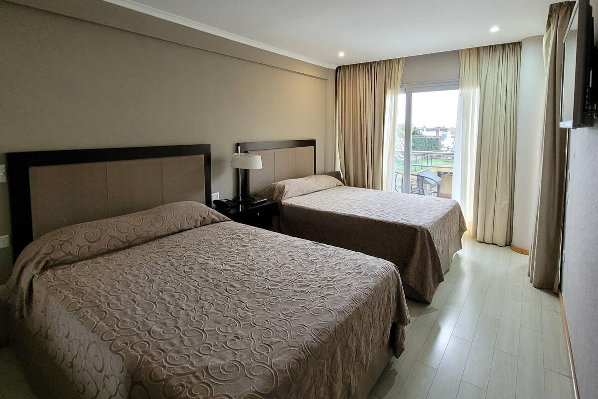 Habitación de hotel con dos camas, una mesita de noche, una lámpara y vistas al exterior.