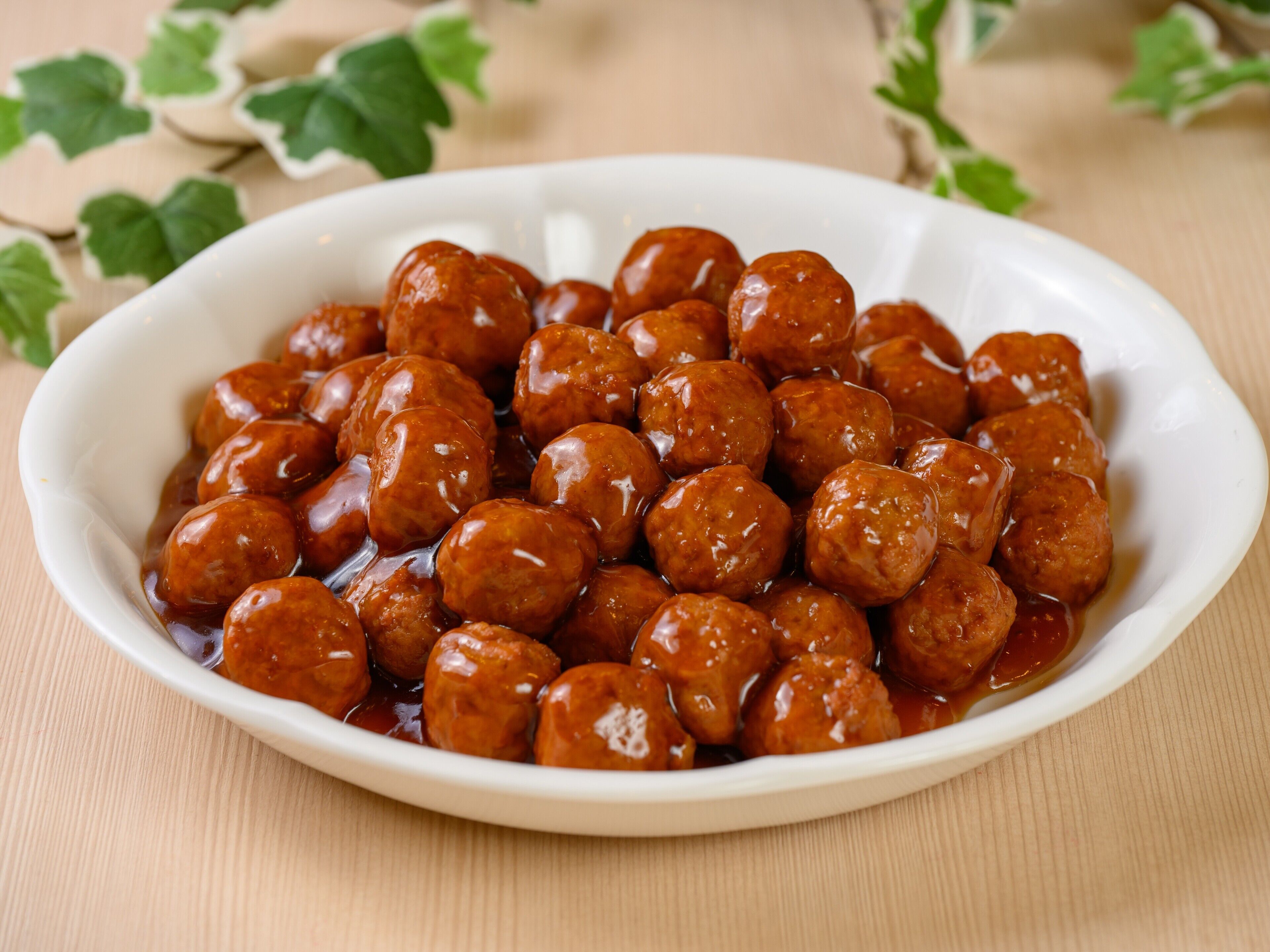 Un plato ovalado blanco lleno de albóndigas redondas y brillantes en salsa, sobre una superficie de madera con hojas verdes al fondo.