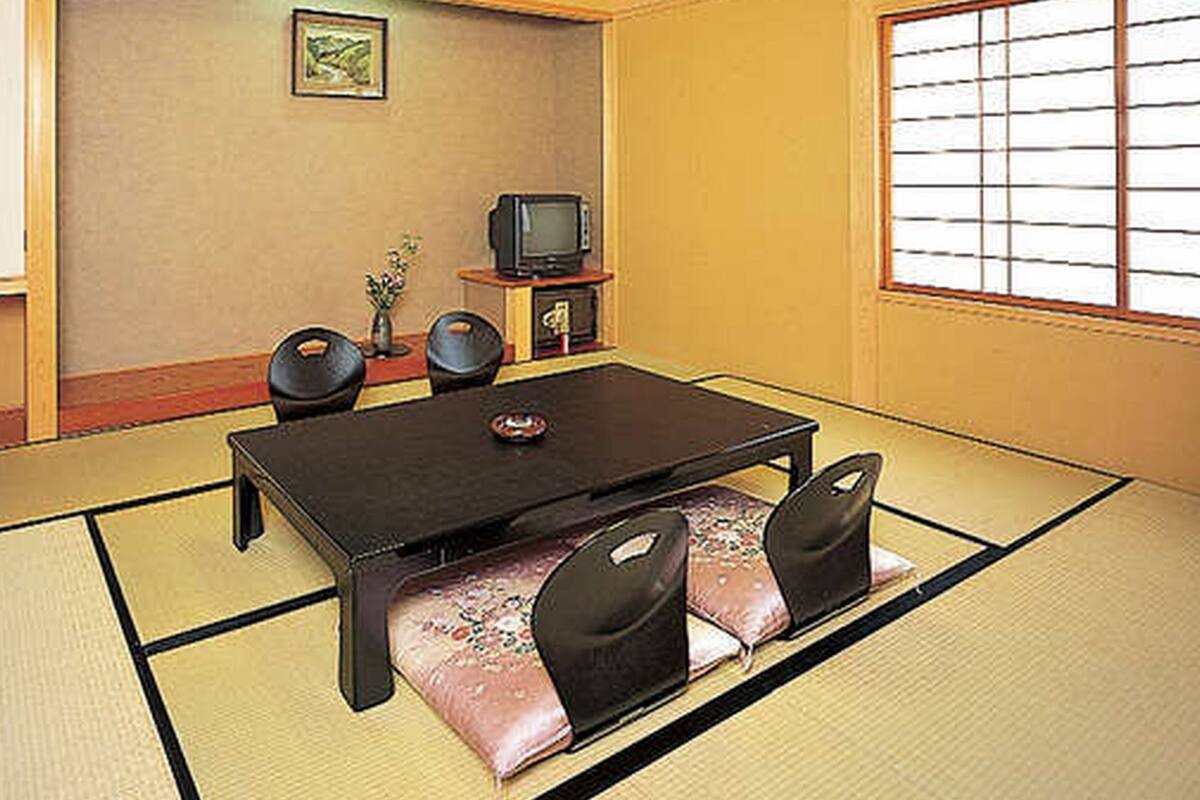 Tatami-mattoilla päällystetty huone, jossa on matala pöytä, tuoleja, televisio ja pieni kaappi.