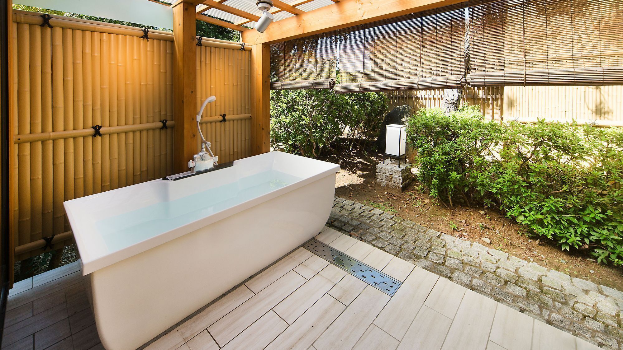 An outdoor bathtub with a wooden pergola overhead, a bamboo fence, and a stone pathway.