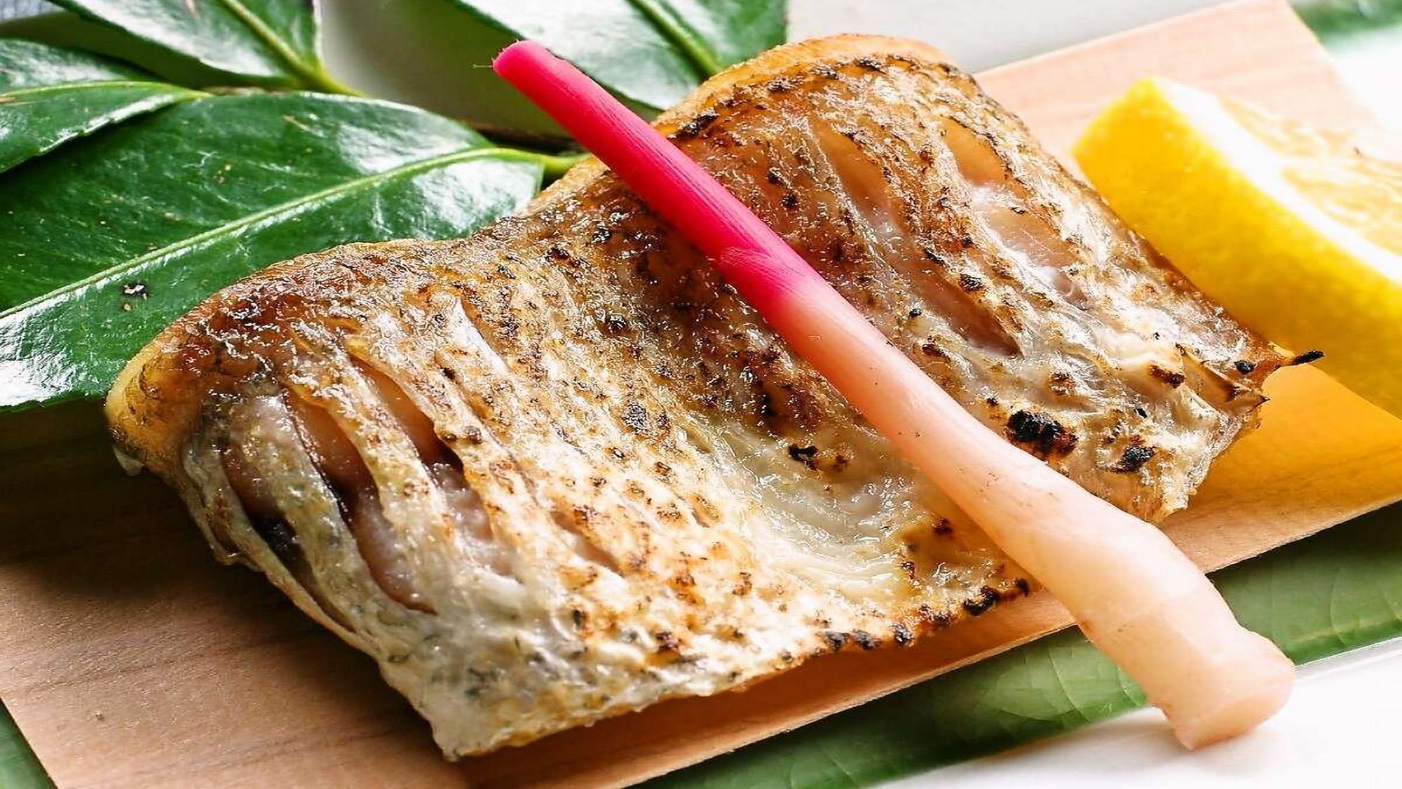 A grilled fish on a wooden board with a lemon wedge and a red straw.