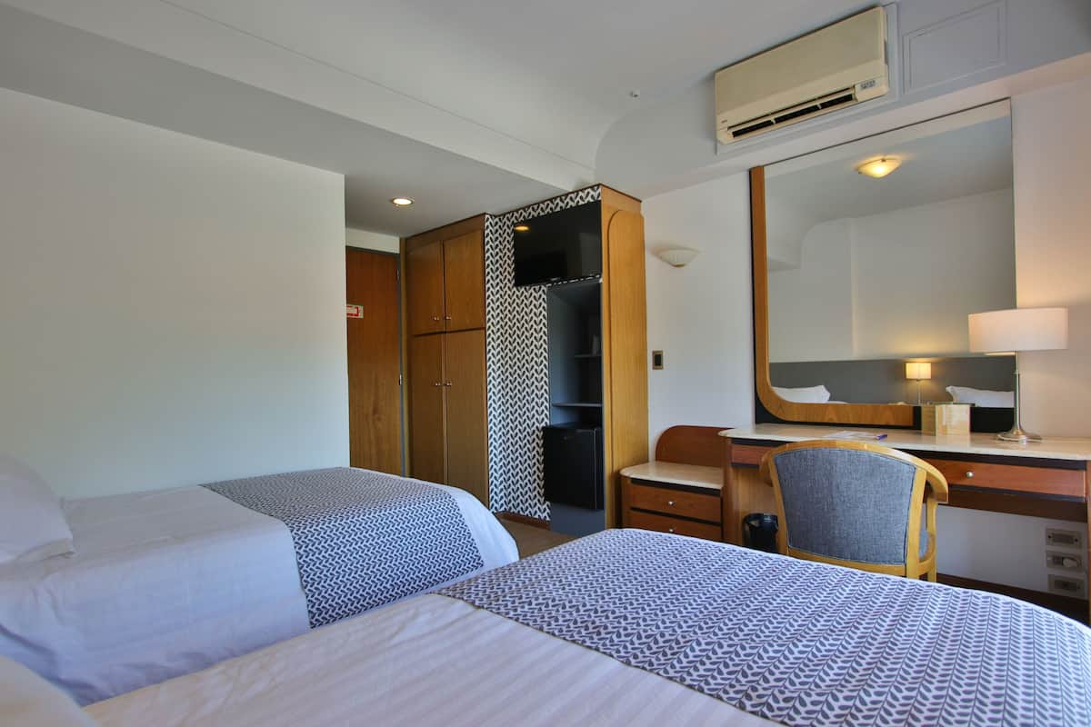 Habitación de hotel con dos camas, un escritorio, una silla, un armario y un espejo.