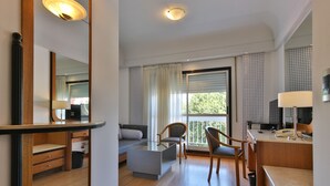 Una habitación de hotel moderna con una mesa de centro de vidrio, un sofá, un escritorio con lámpara y un ventanal con cortinas.