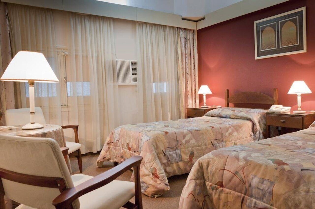 A hotel room with two beds, a desk, a chair, a lamp, and a wall-mounted phone.