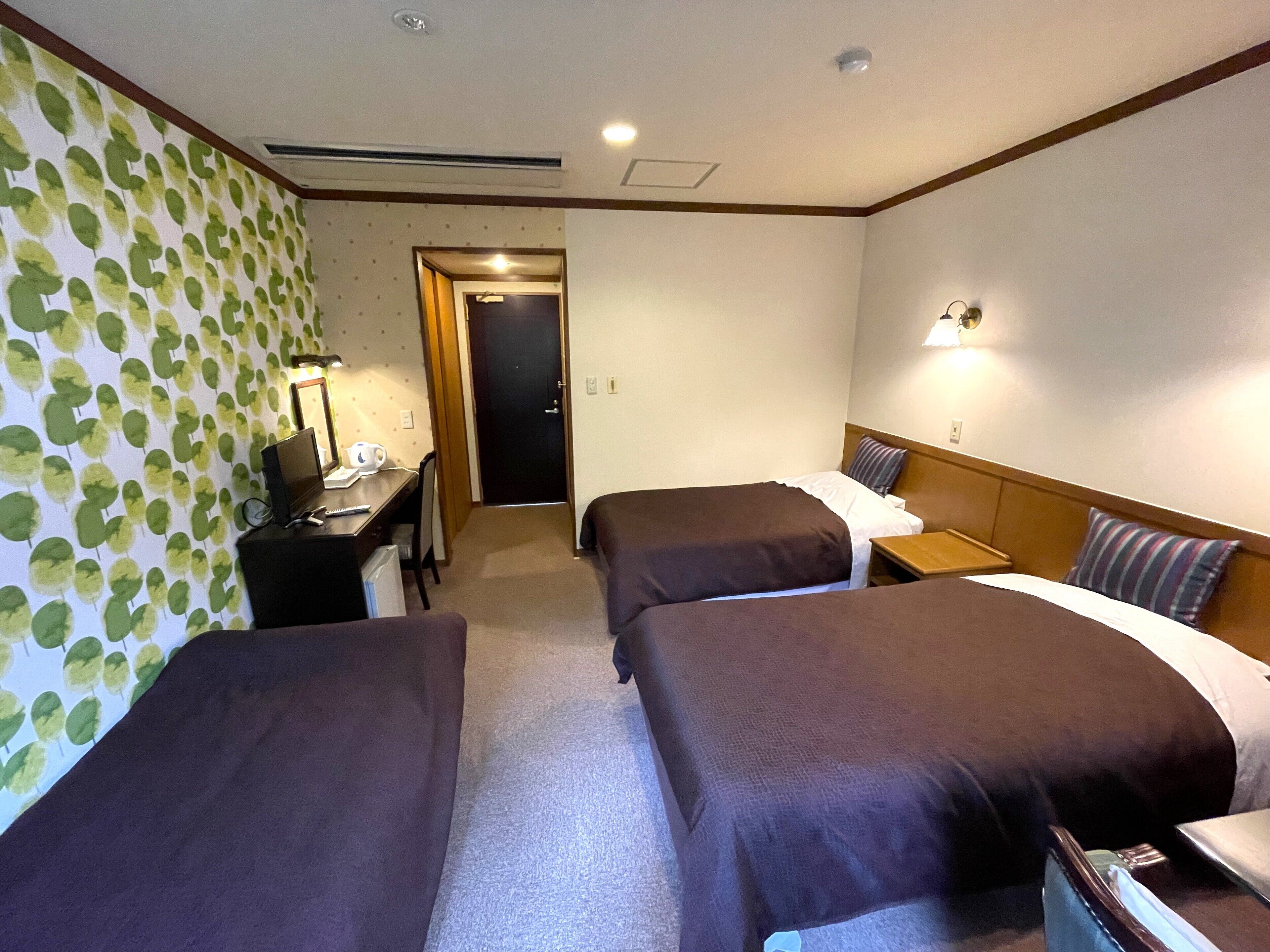 Ein Hotelzimmer mit zwei Einzelbetten, einem Schreibtisch mit Stuhl, einer Lampe und einer Tür zu einem weiteren Raum.